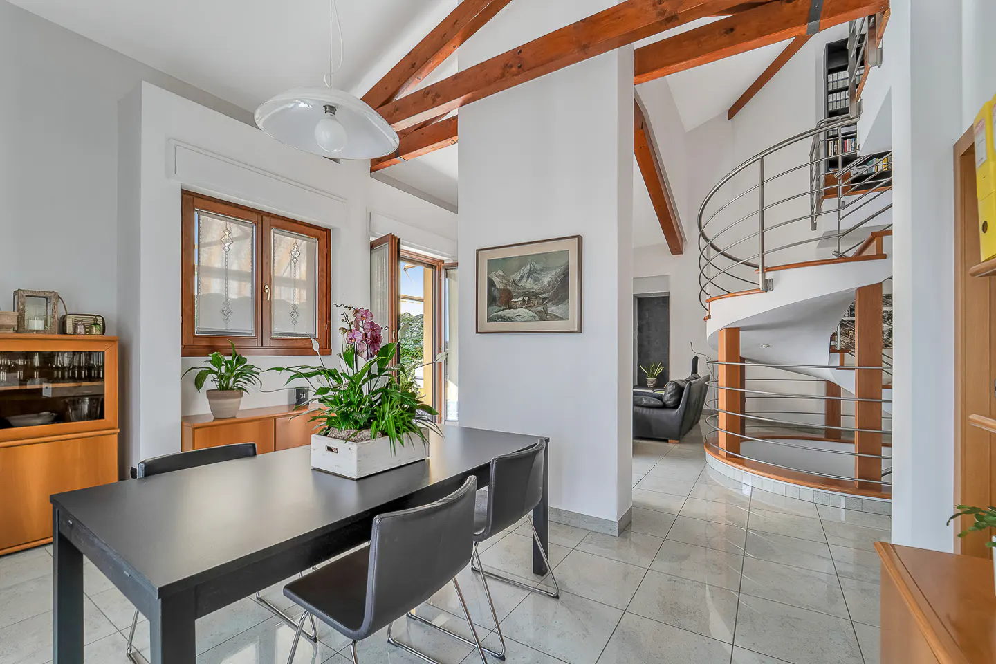 Bright, modern dining room with a black table, chairs, and a spiral staircase in the background. Wooden beams accent the white walls.