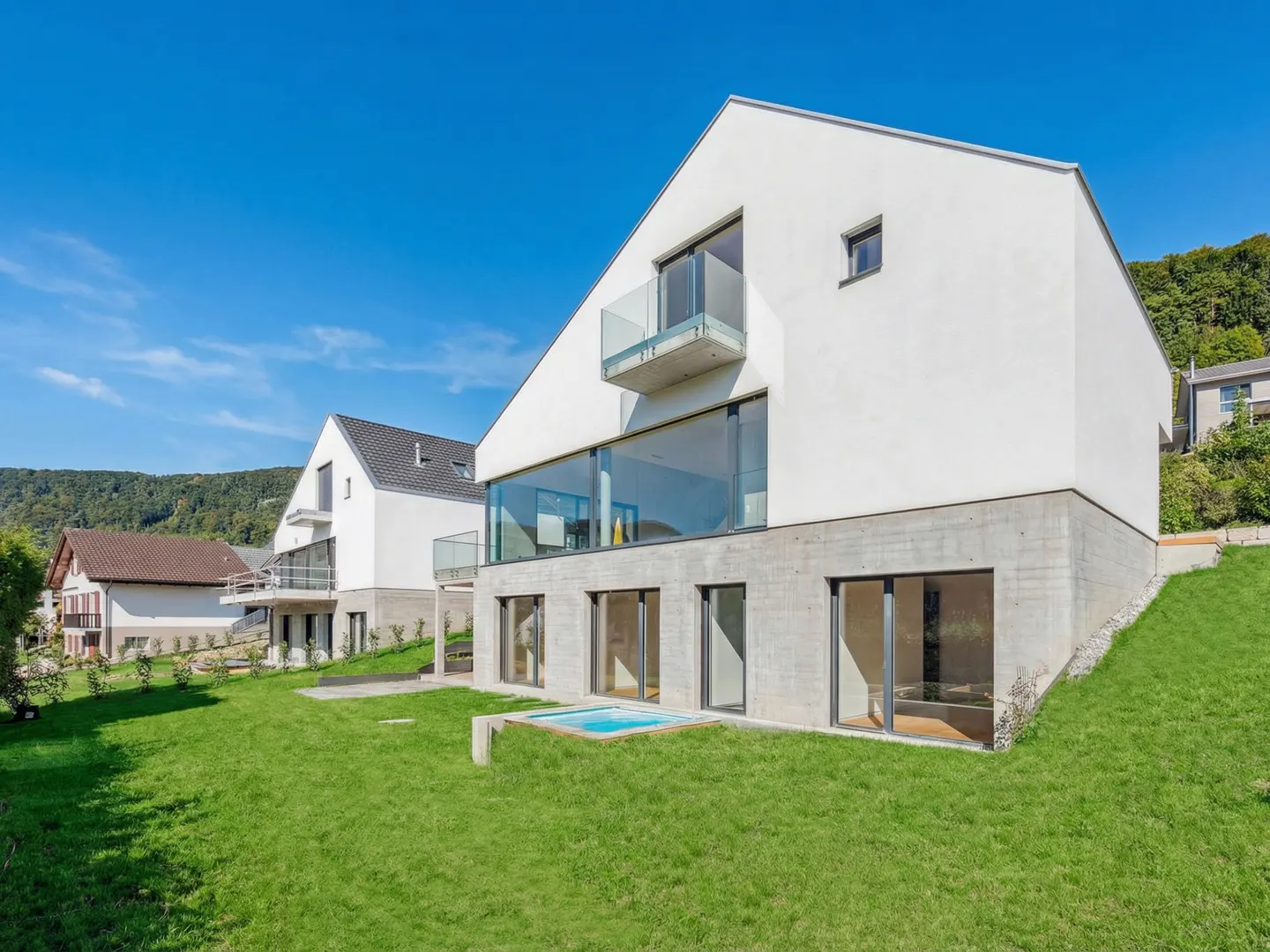 Modern white house with glass windows and balcony on a green lawn under a blue sky.
