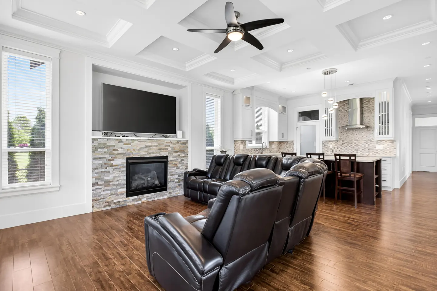 Bright, open-concept living room with dark wood floors, black leather recliners, stone fireplace, and white kitchen in the background.