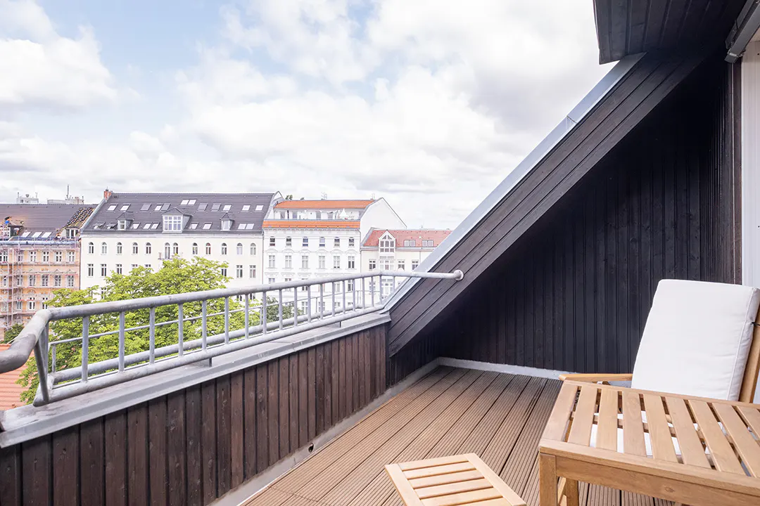 Balcony with wood flooring, metal railing, and dark wood siding. A chair and table sit on the balcony overlooking city buildings.