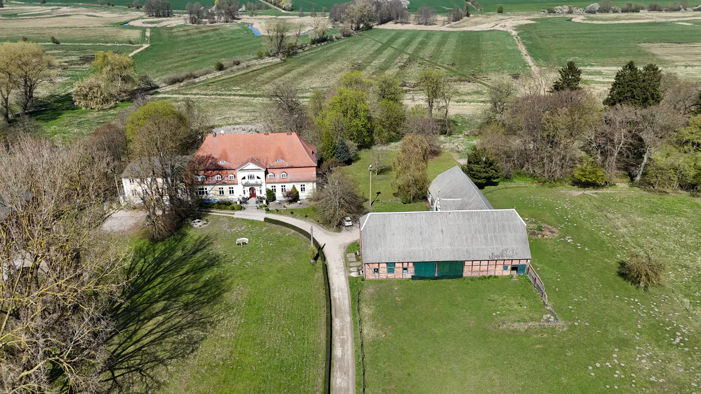 Aerial view of a white manor house with a red tile roof, a barn, and green fields.
