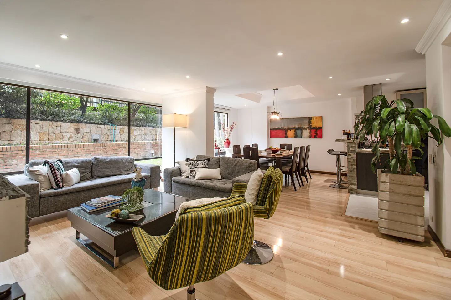 Bright, open-concept living space with gray sofas, striped chairs, and a dining area. Large windows offer a view of a stone wall and greenery.