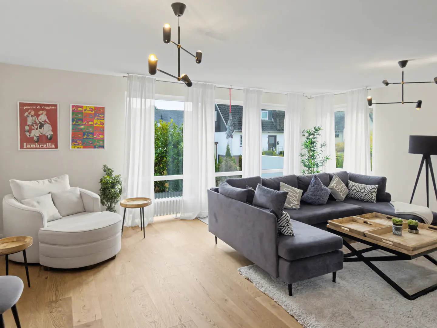 Bright living room with a gray sectional sofa, white armchair, wood floors, and modern lighting fixtures. Large windows with white curtains.