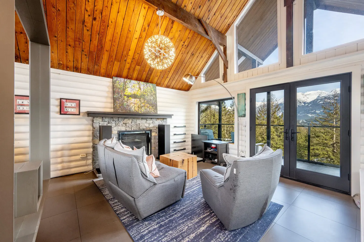 Living room with wood ceiling, stone fireplace, gray sofas, and mountain view through black framed glass doors.