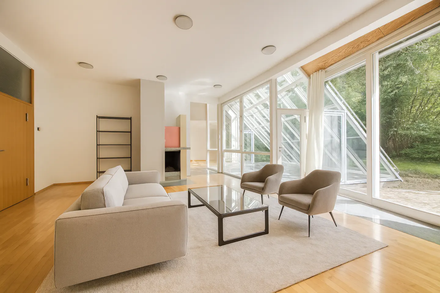 Bright living room with a beige sofa, two chairs, and a glass coffee table on a white rug. Large windows overlook green trees.
