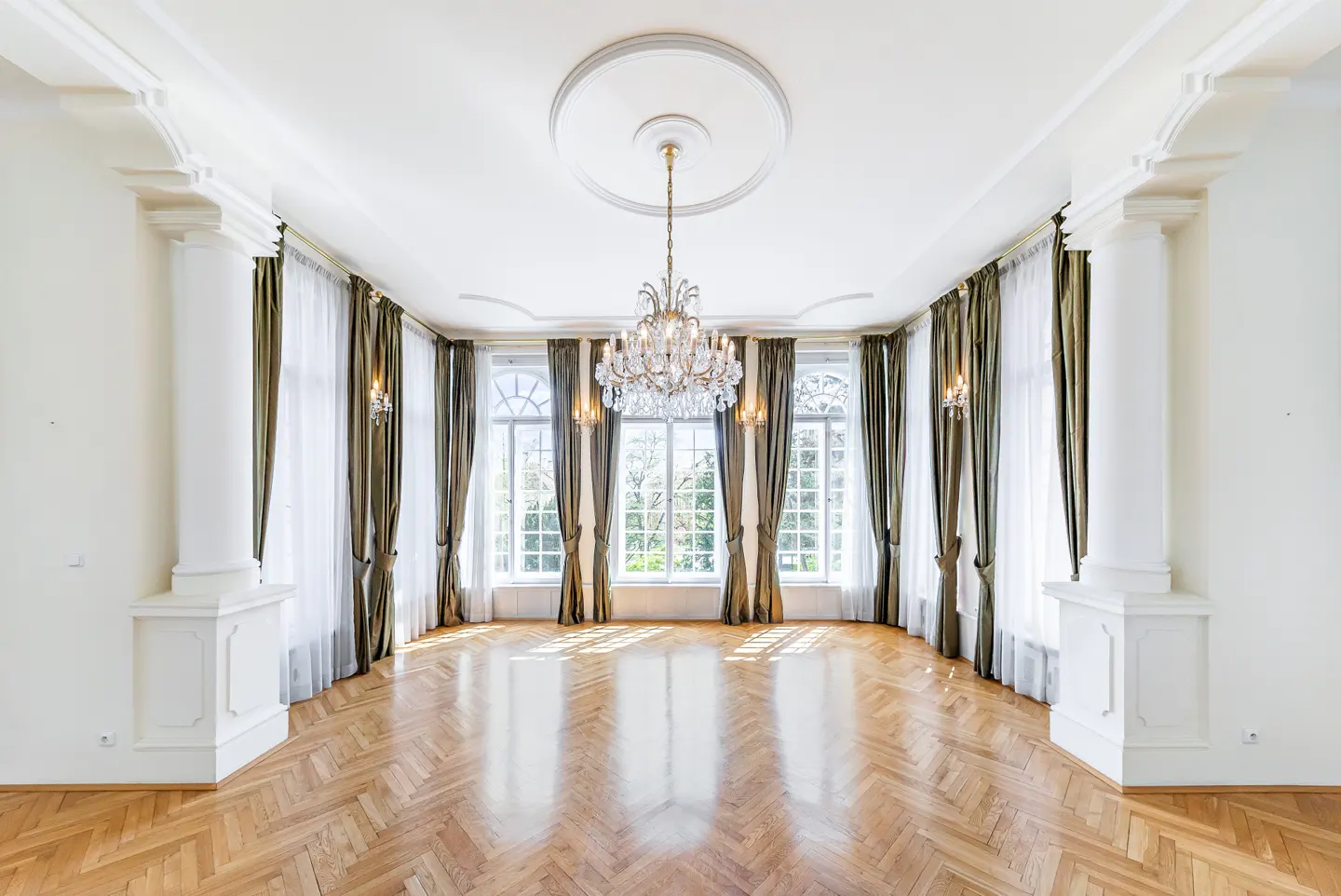 Bright, empty room with herringbone wood floors, white walls, columns, and a crystal chandelier. Windows with green curtains let in natural light.