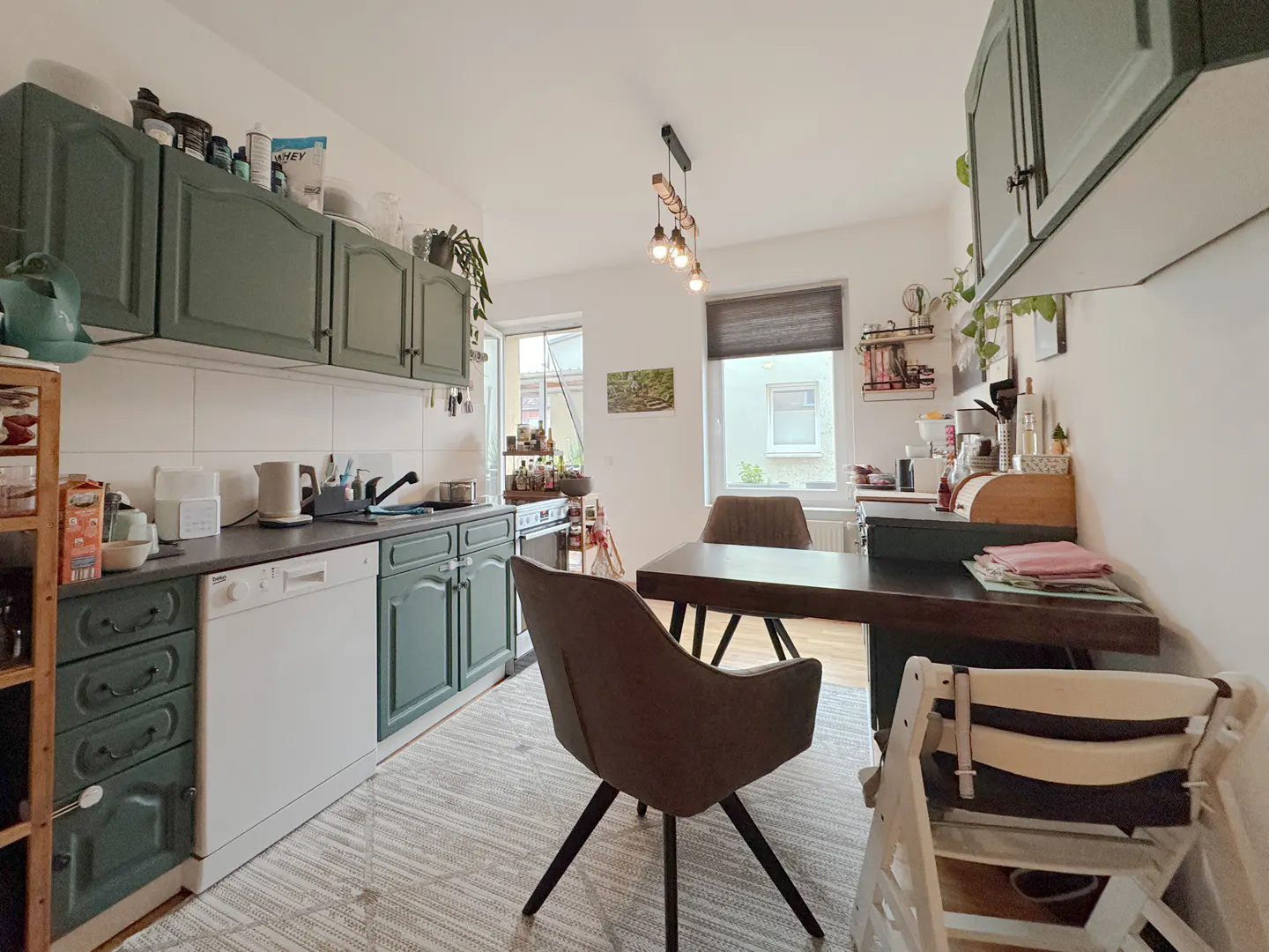 A kitchen with green cabinets, a white dishwasher, and a dark wood table with brown chairs. A high chair is near the table.