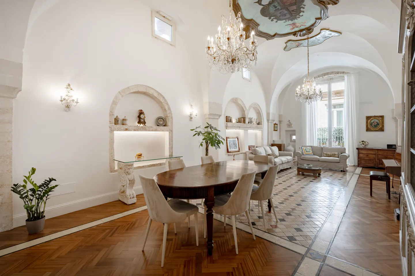 A bright, elegant dining and living room with a wood table, chandeliers, and arched niches.