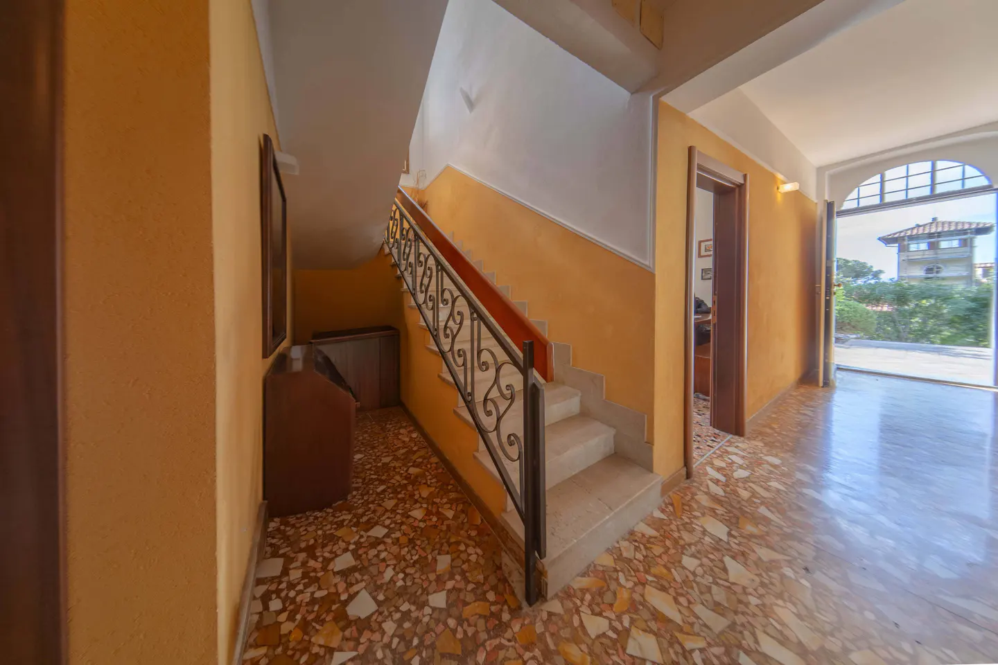 Foyer with orange walls, a staircase with a wrought iron railing, and a doorway leading to an outdoor view of a building.