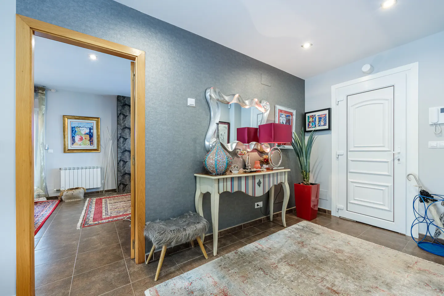 Foyer with gray walls, brown tile floor, and a console table with a silver mirror and red lamps. A white door is on the right.