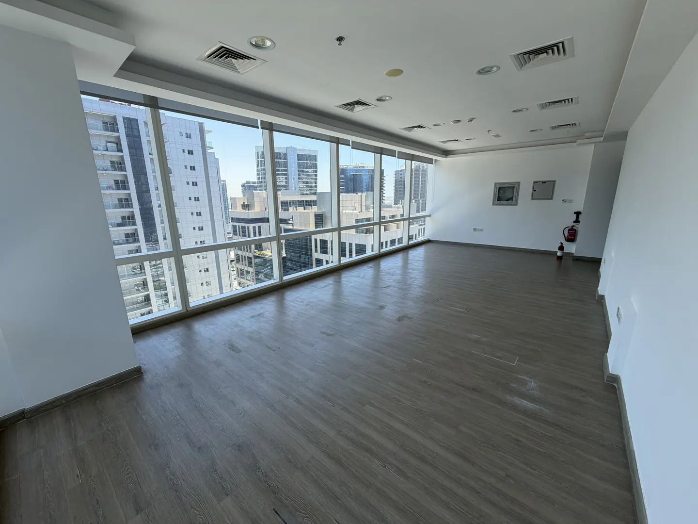 Empty office space with wood-look flooring, white walls, and floor-to-ceiling windows overlooking a city skyline.
