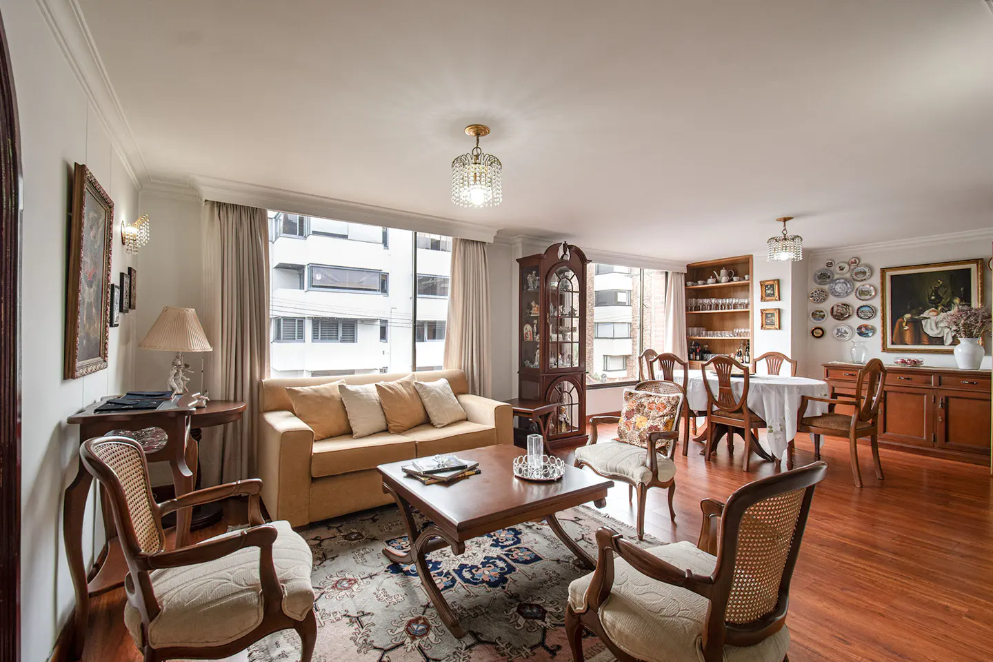 Bright living and dining room with hardwood floors, a tan sofa, and a wooden dining set. Large windows offer city views.