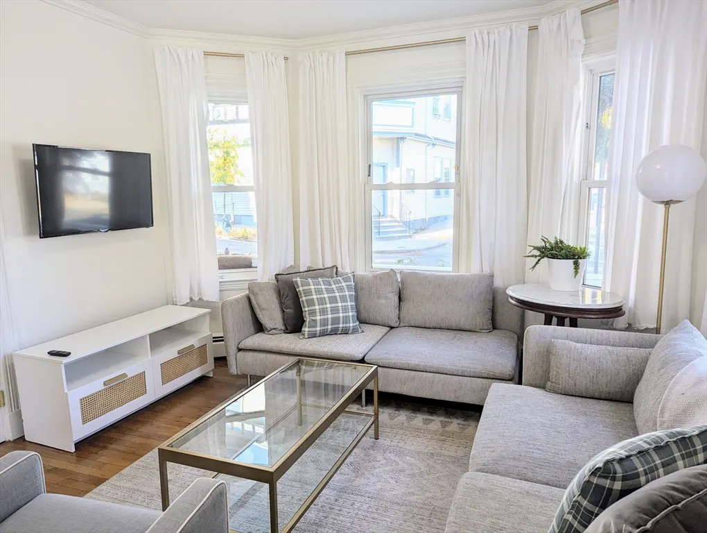 Bright living room with gray sectional sofas, a glass coffee table, white media console, and windows with white curtains.
