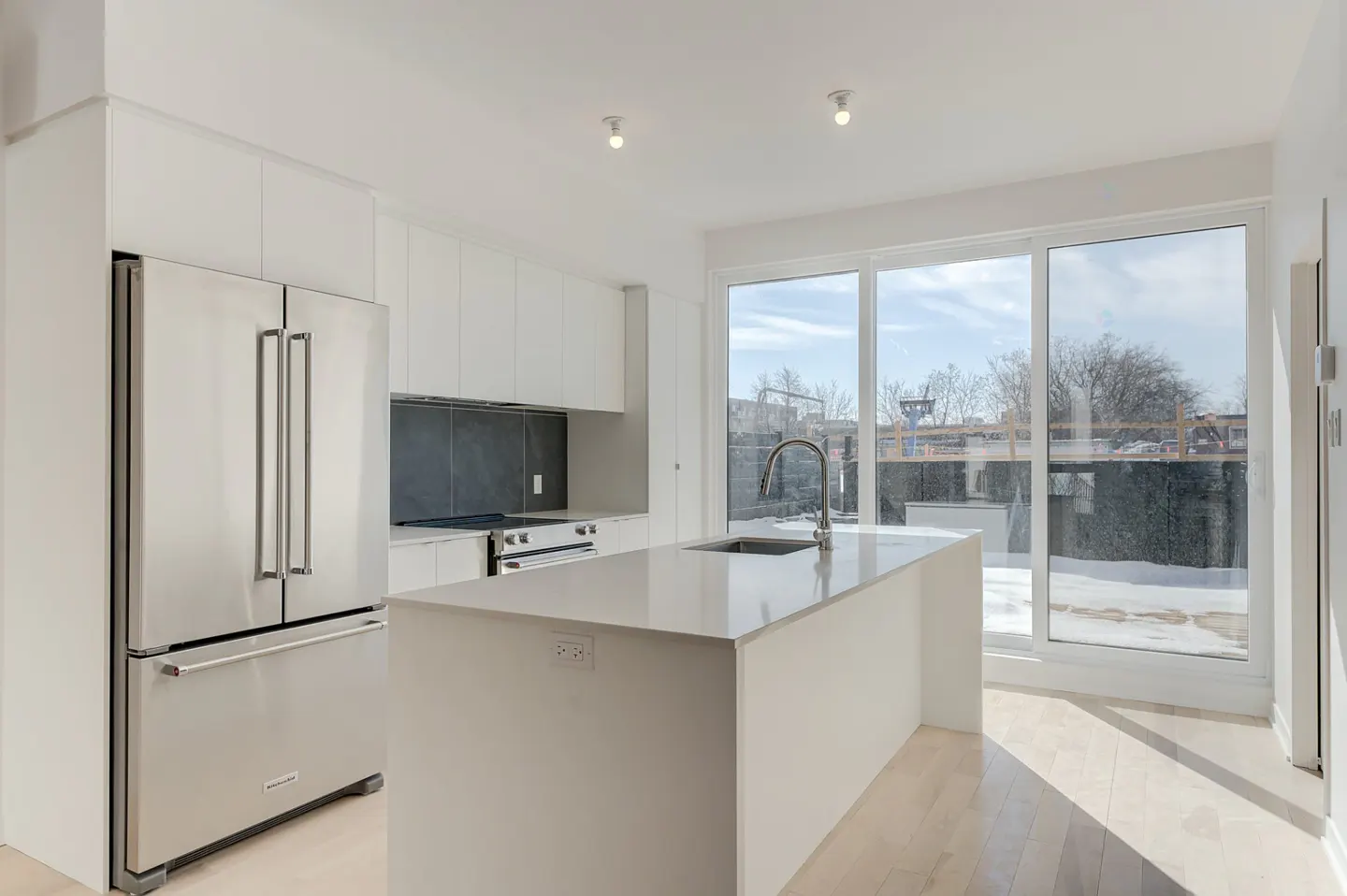 Bright, modern kitchen with white cabinets, stainless steel fridge, and island. Large windows overlook a snowy backyard.