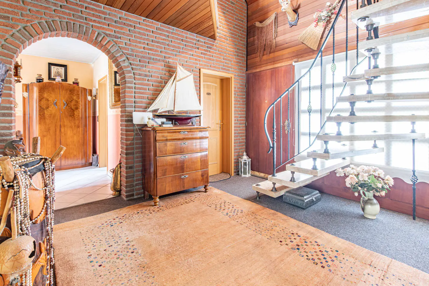 Interior view of a home with a brick archway, wooden dresser with a sailboat model, and a spiral staircase with white steps.