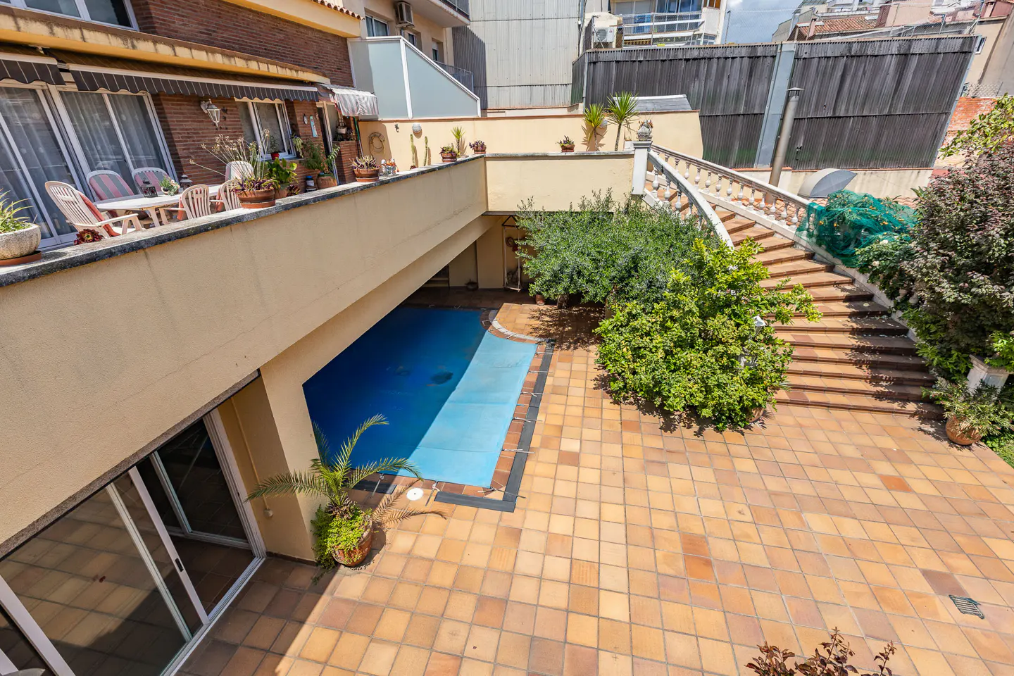 Overhead view of a home's outdoor space with a blue-covered pool, tiled patio, and a staircase with stone railings. Balcony with furniture and plants above.
