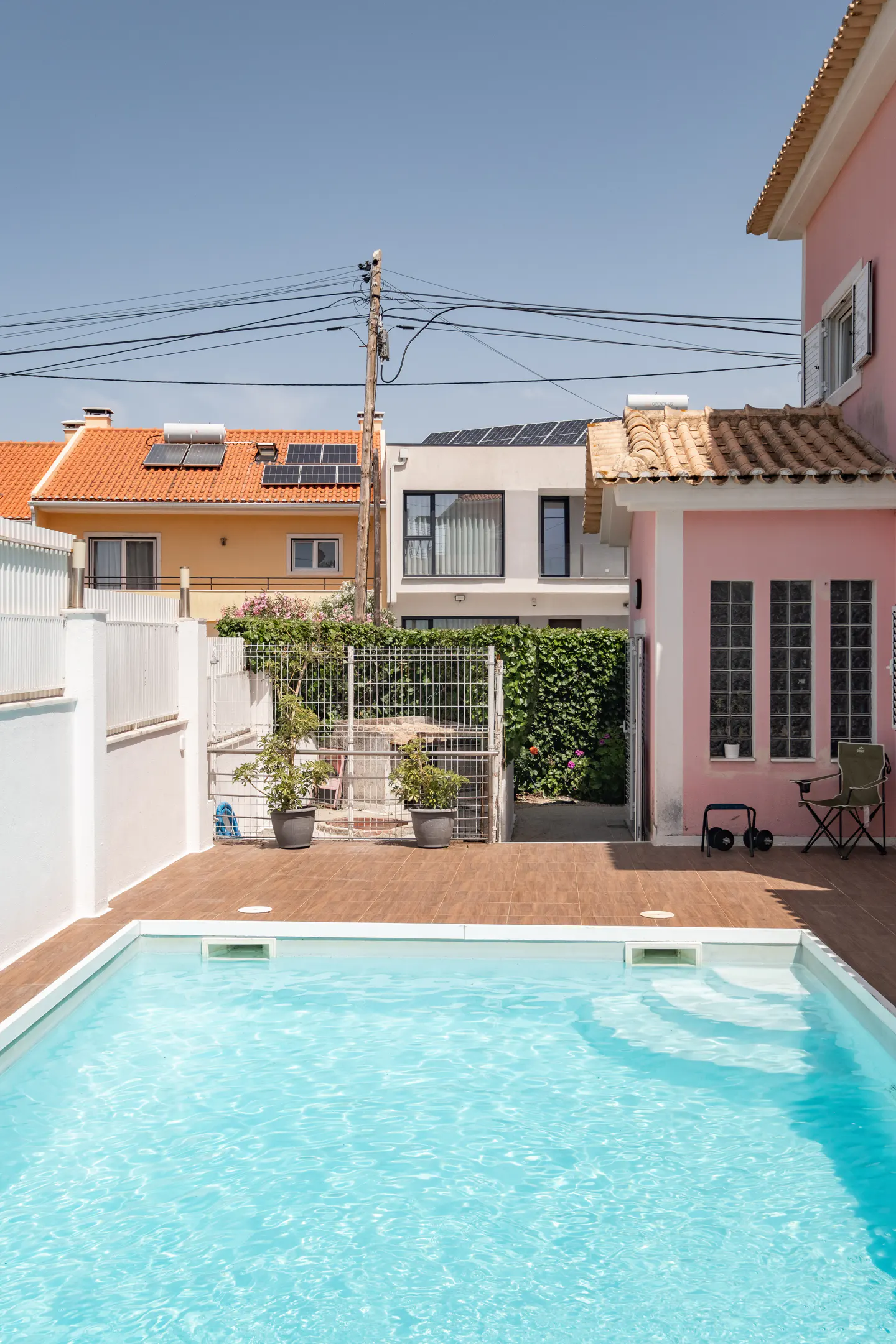 A backyard pool with clear blue water, surrounded by a brown tiled deck and a pink house.