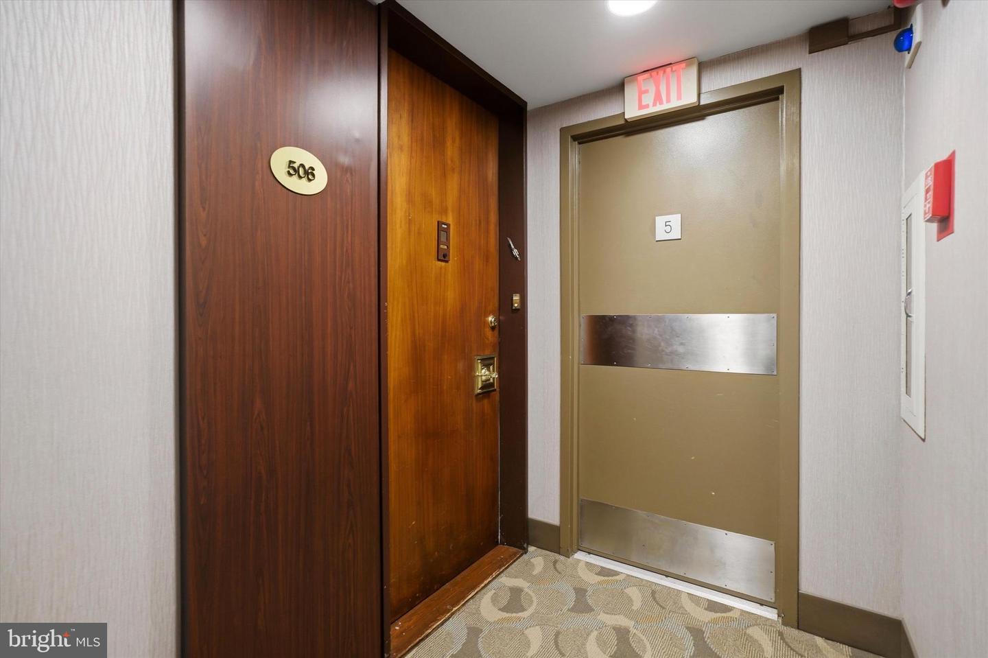 Hallway view with apartment door 506, exit door 5, and fire alarm. The walls are white and the carpet is patterned.
