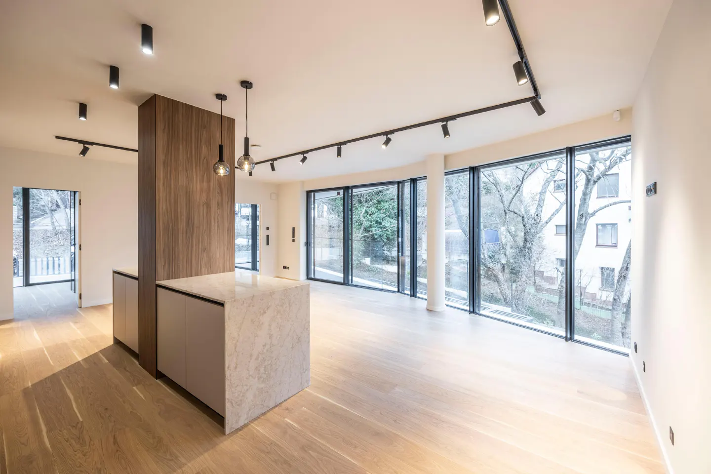 Bright, modern open-plan living space with wood floors, a marble kitchen island, and large windows overlooking trees. Track lighting on the ceiling.