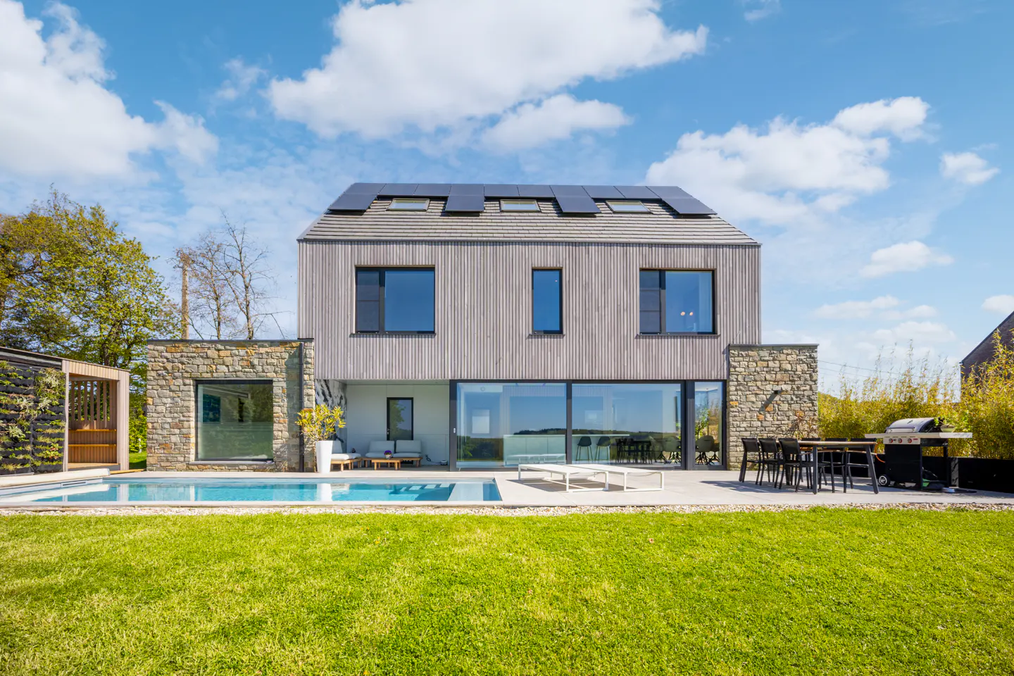 Modern home with a pool and green lawn under a blue sky. The house has wood siding, stone accents, and solar panels on the roof.