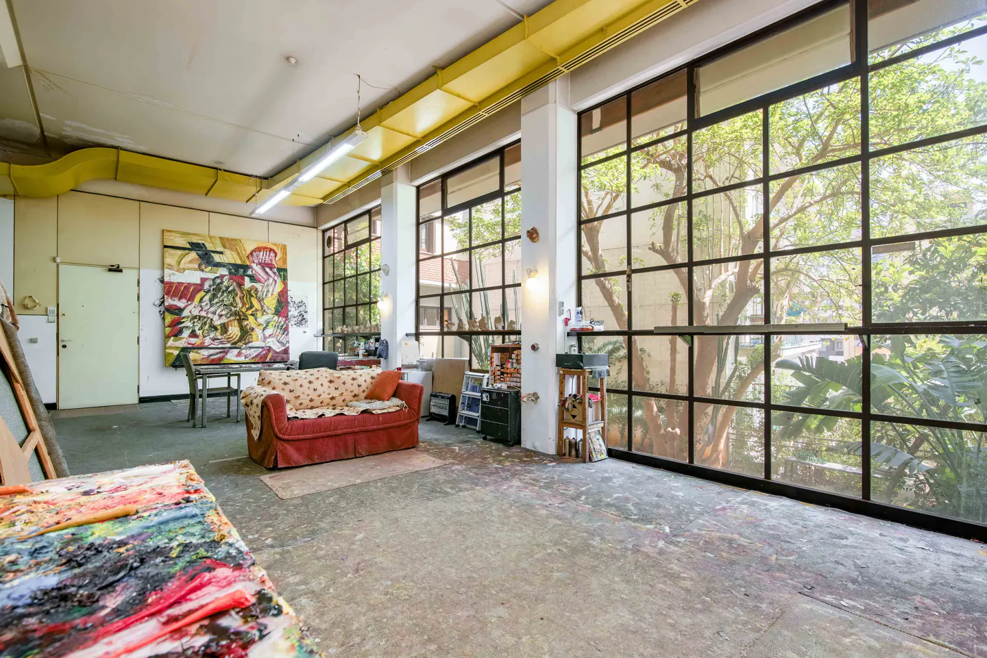 Bright, airy studio with large windows, a red couch, and abstract art. Yellow ductwork runs along the ceiling.