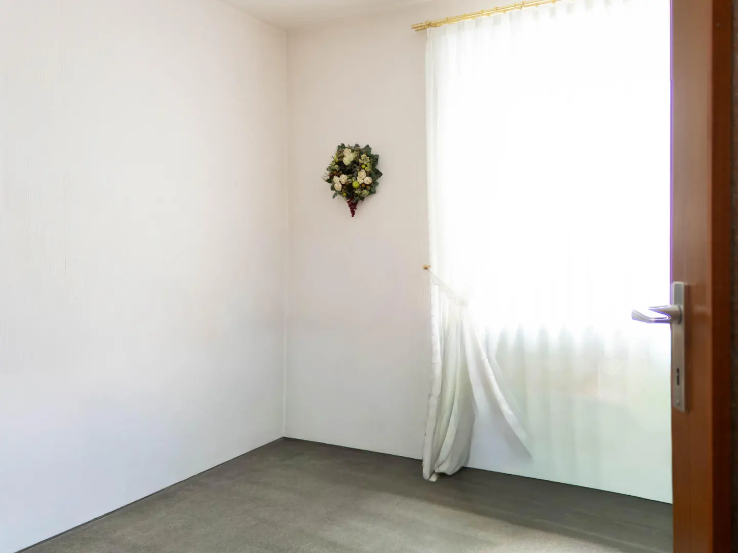 Bright, empty room with white walls, gray carpet, and an open wood door. A floral wreath hangs on the wall near a window with sheer white curtains.
