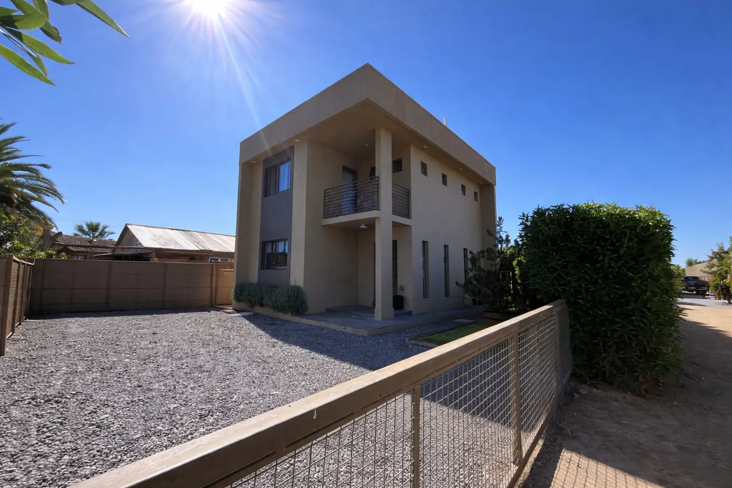 Two-story beige house with a small balcony, gravel driveway, and a metal fence under a clear blue sky.