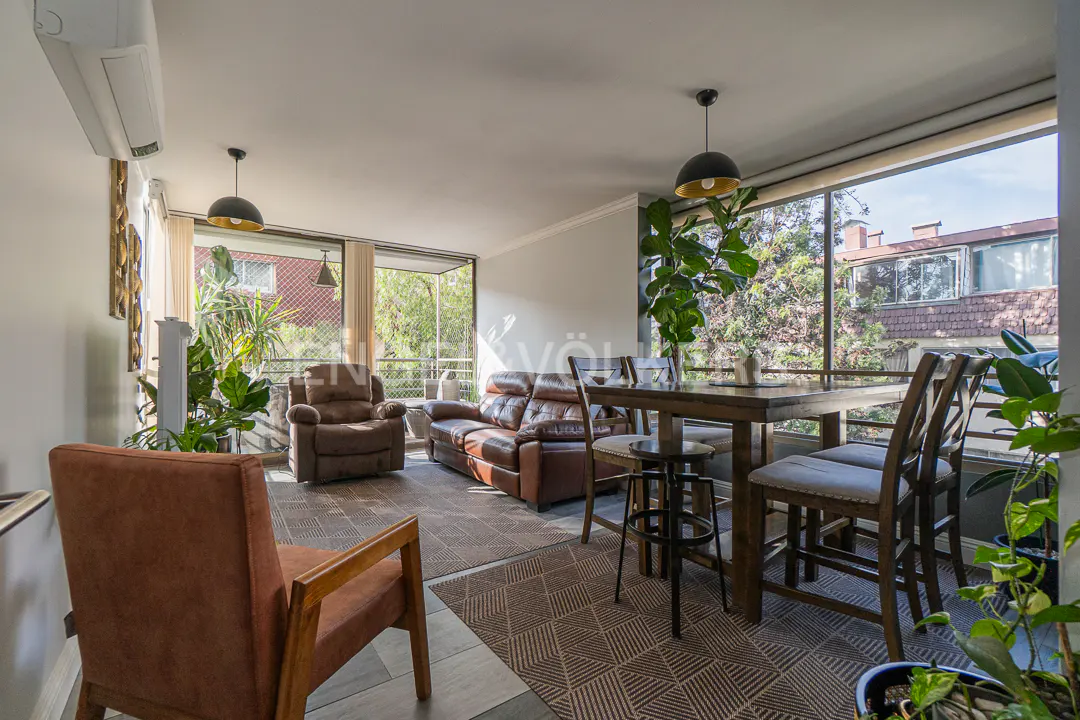 Bright living and dining area with brown leather sofa, armchair, wooden dining set, and large windows overlooking trees.