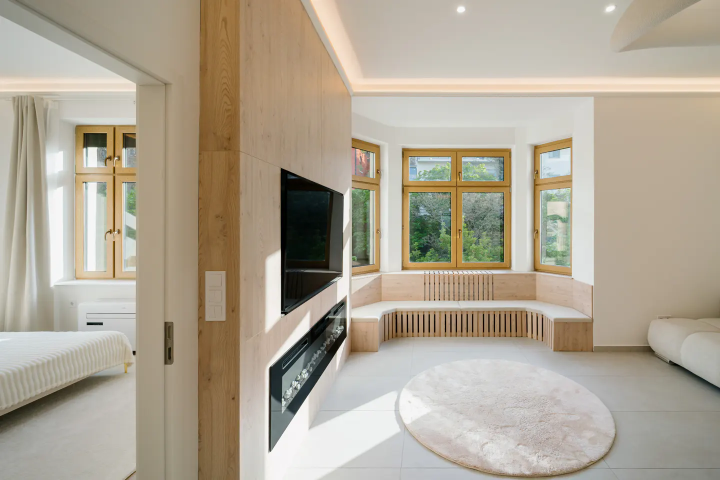Bright living room with a bay window, a built-in bench, and a round rug. A TV and fireplace are mounted on a wood-paneled wall. A bedroom is visible through a doorway.