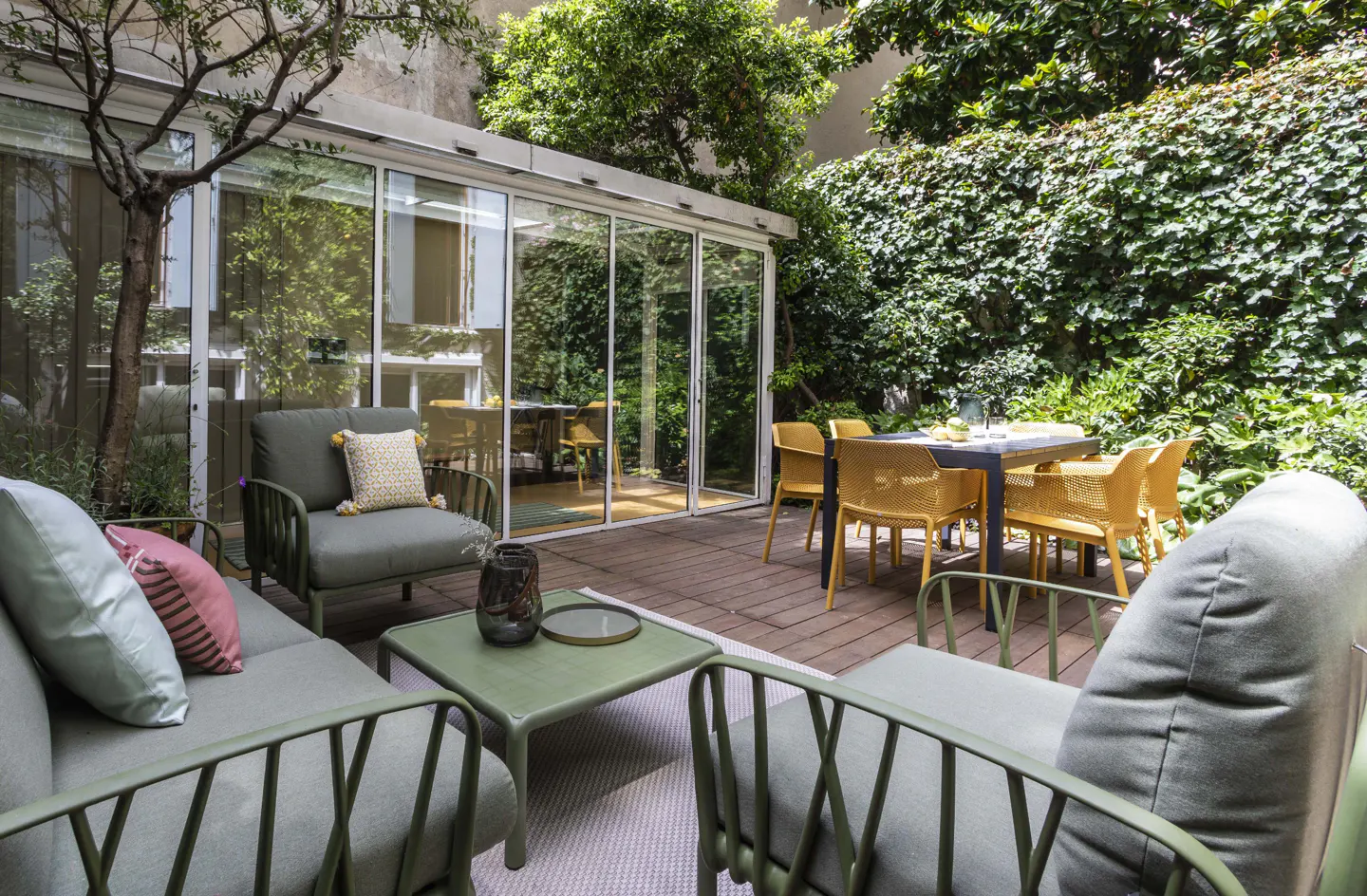 Outdoor patio with green furniture, a glass-enclosed room, and a dining table with yellow chairs. Green foliage surrounds the space.
