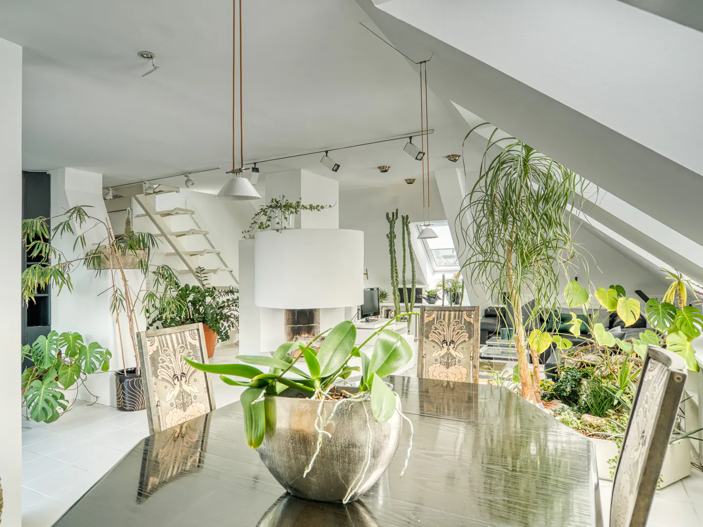 Bright, modern dining room with a dark wood table, chairs, and many plants. A white fireplace and stairs are in the background.
