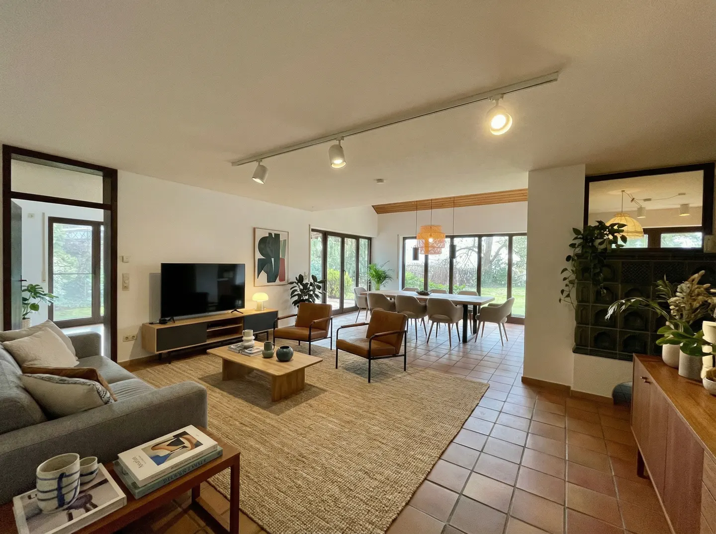 Open-concept living and dining area with a gray sofa, brown leather chairs, and a dining table with chairs. Large windows offer outdoor views.