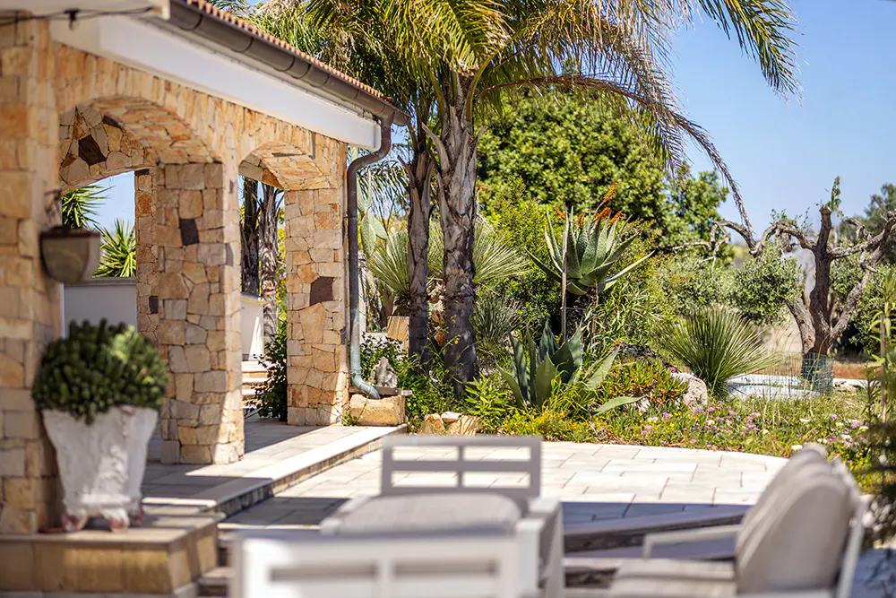 Stone archway leads to a patio with white furniture, surrounded by lush greenery and a palm tree under a blue sky.