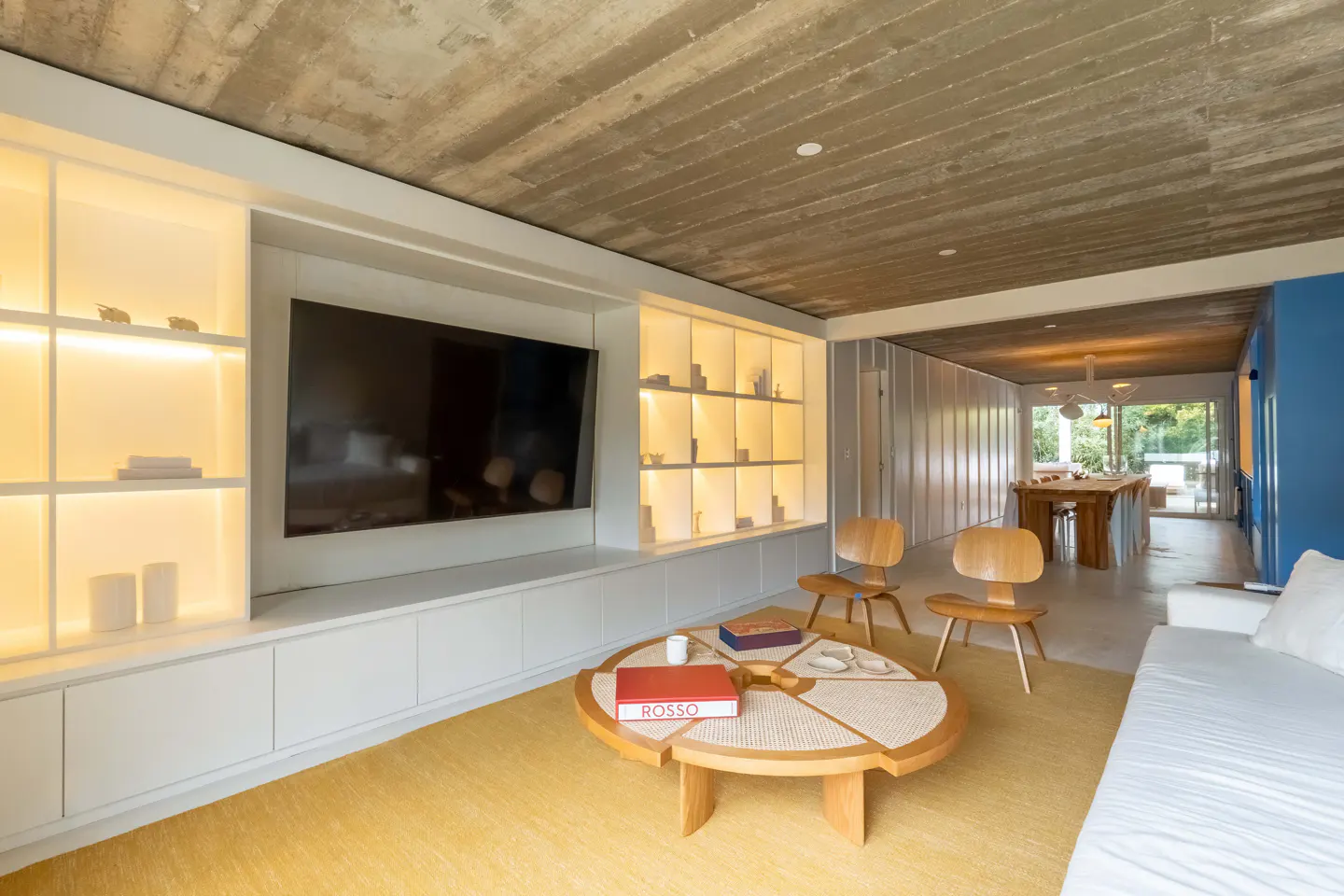 Bright living room with a yellow rug, a round wooden table, and two wooden chairs. A large TV is mounted above a white cabinet with illuminated shelves.