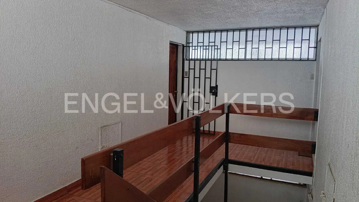 Interior view of a staircase with wood railings and tile floors, leading to a barred window and door. The walls are textured and painted white.