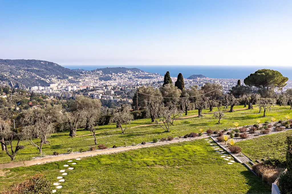 Scenic view of Nice, France, featuring olive trees, a green lawn, and a stone path leading to the city and the Mediterranean Sea.
