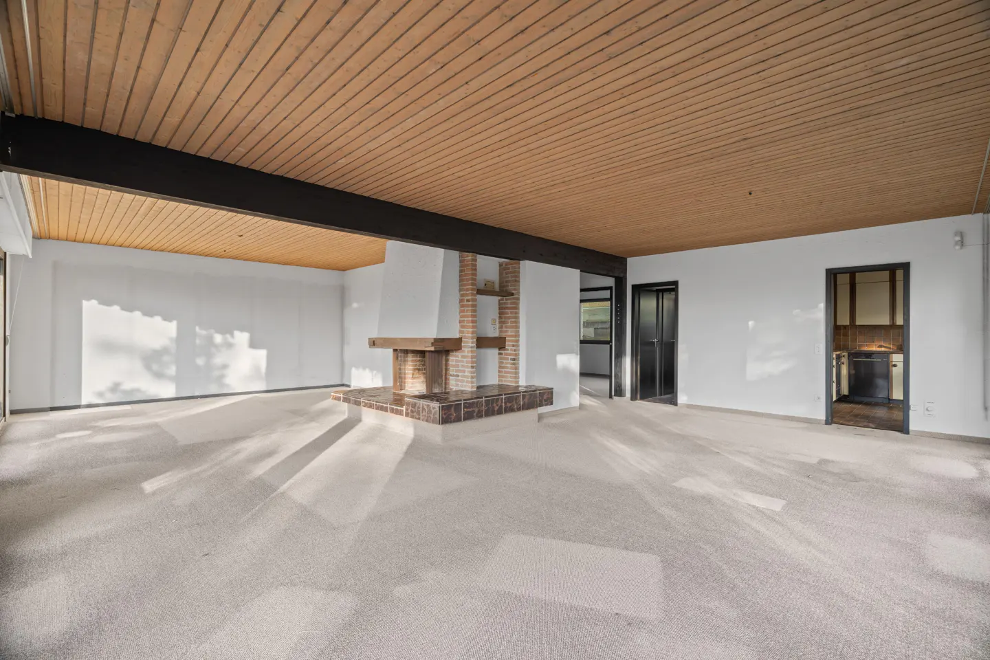 An empty living room with white walls, a wood ceiling, and a brick fireplace. Sunlight streams in from the left.