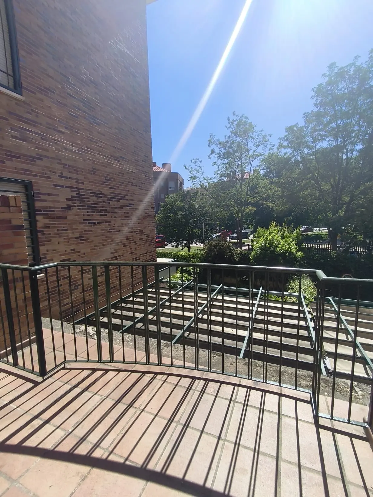 A sunny balcony with a tiled floor, dark metal railing, and a view of a brick building and green trees.