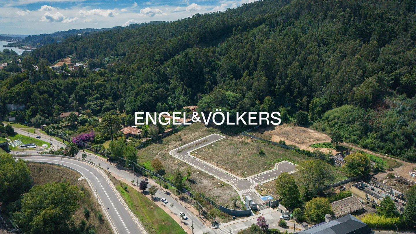 Aerial view of a property lot with roads, surrounded by lush green trees and hills. Engel & Völkers logo overlay.