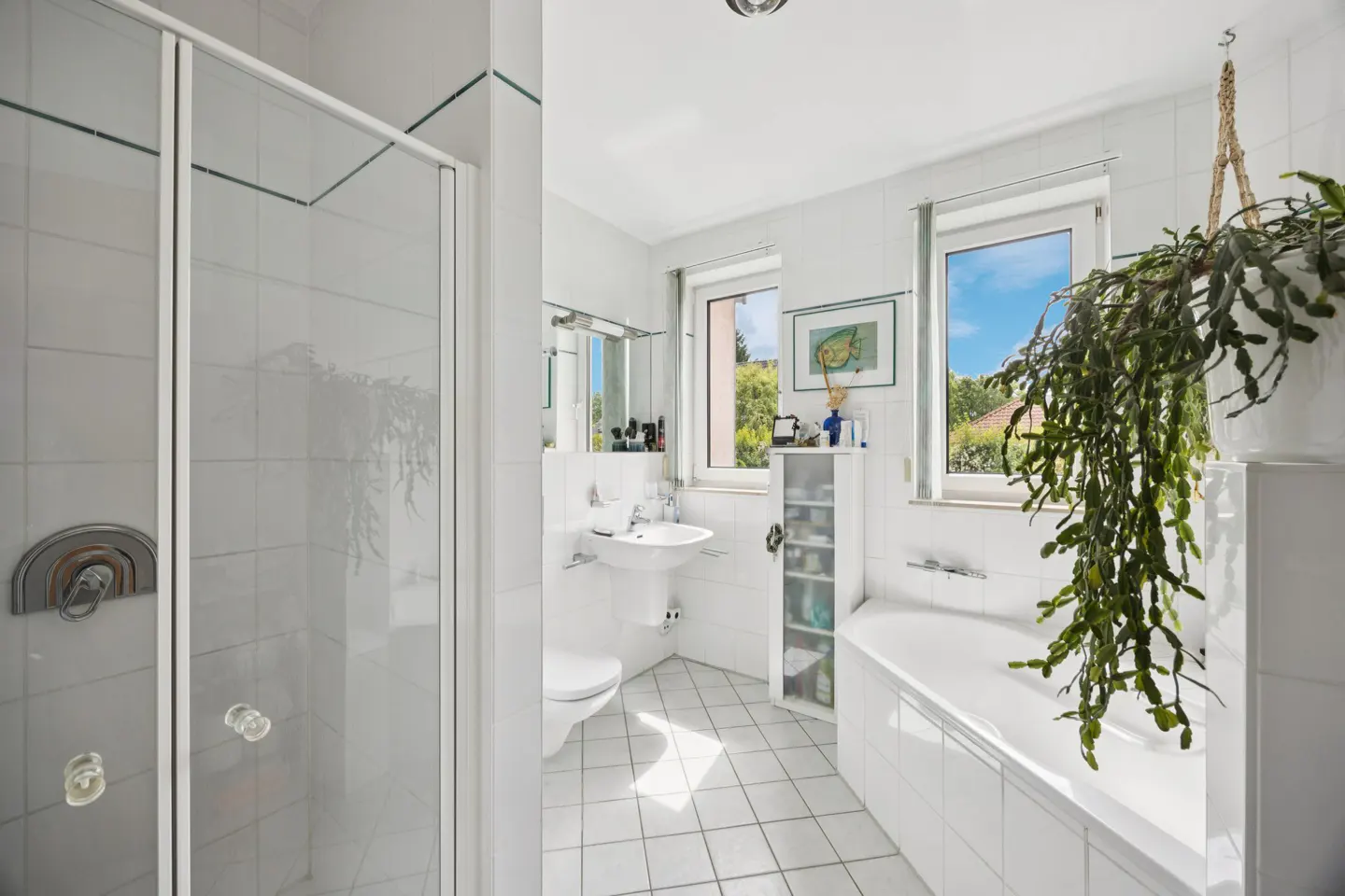 Bright white bathroom with a glass shower, pedestal sink, toilet, bathtub, and a large hanging plant by the window.