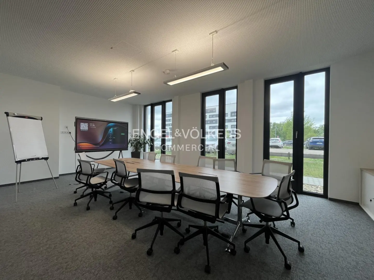 Bright conference room with a long wooden table, black chairs, a whiteboard, and a large screen. Windows overlook a parking lot.