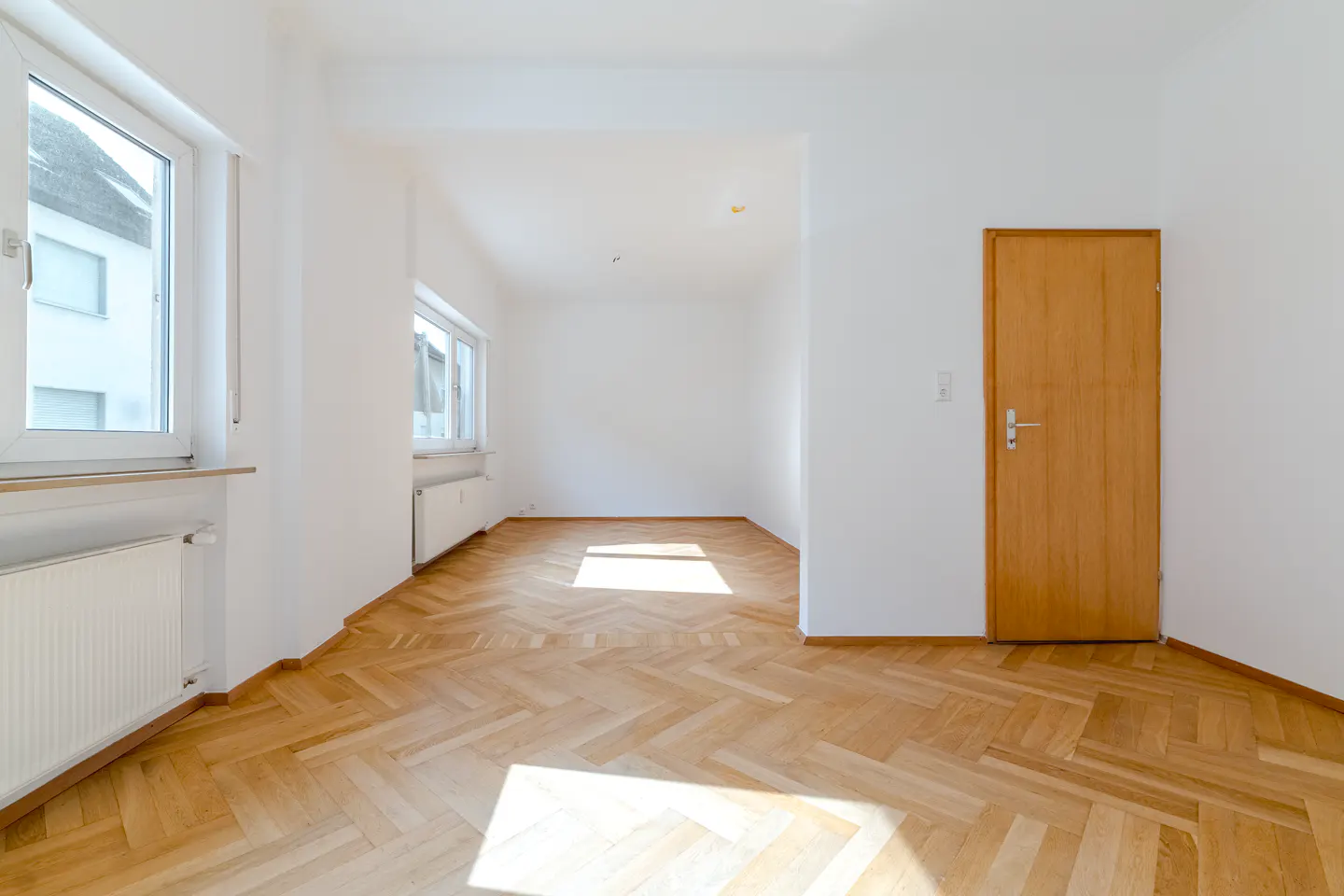 Bright, empty room with herringbone wood floors, white walls, windows, and a light wood door.