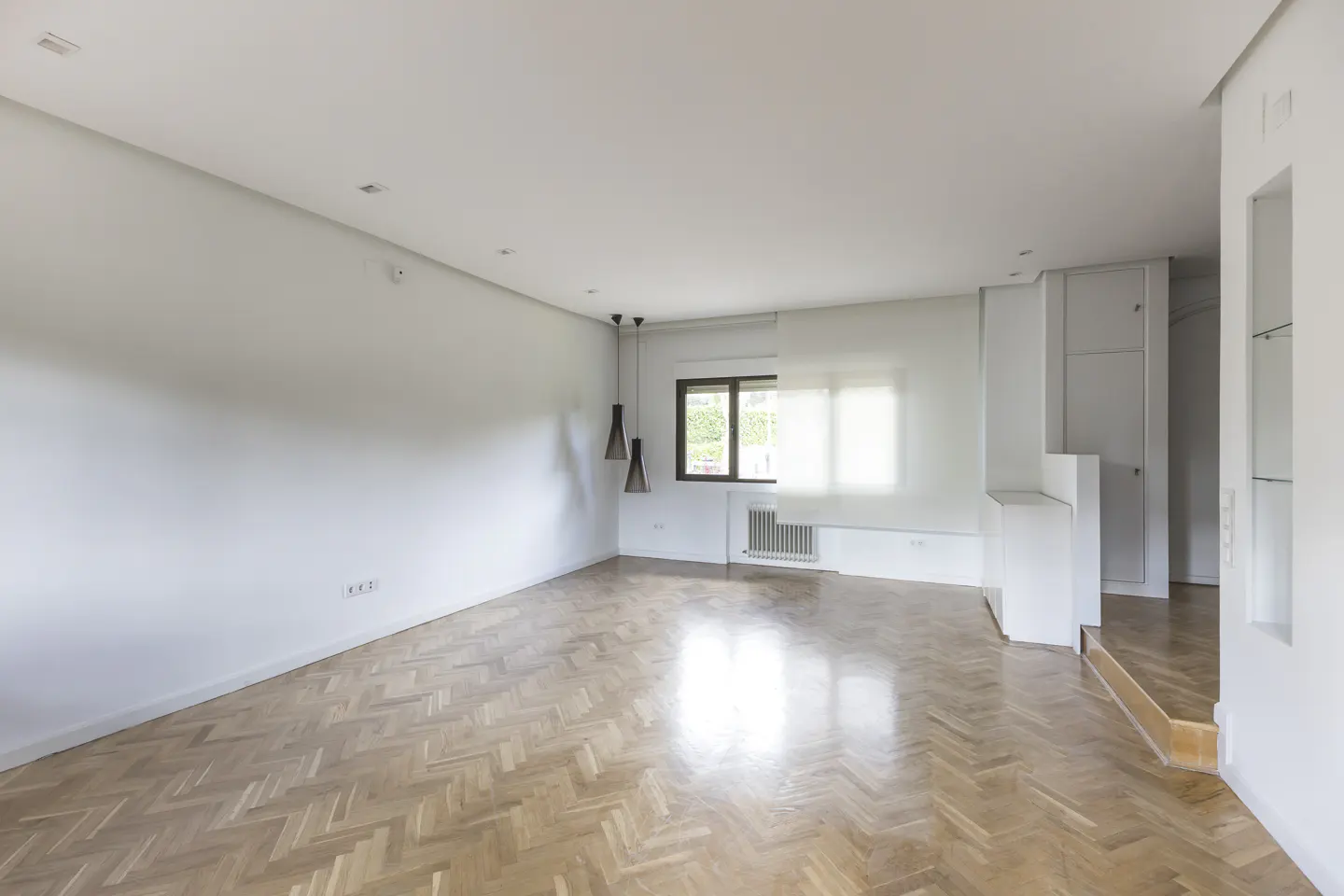 Bright, empty room with herringbone wood floors, white walls, and a window with sheer curtains. Three pendant lights hang from the ceiling.
