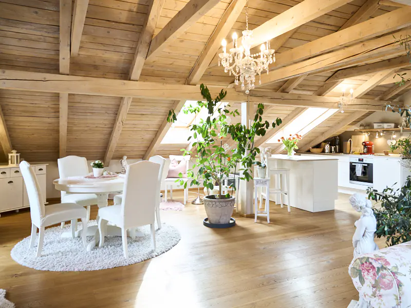 Attic apartment with exposed wood beams, chandelier, dining table, and kitchen. The room is bright and airy.