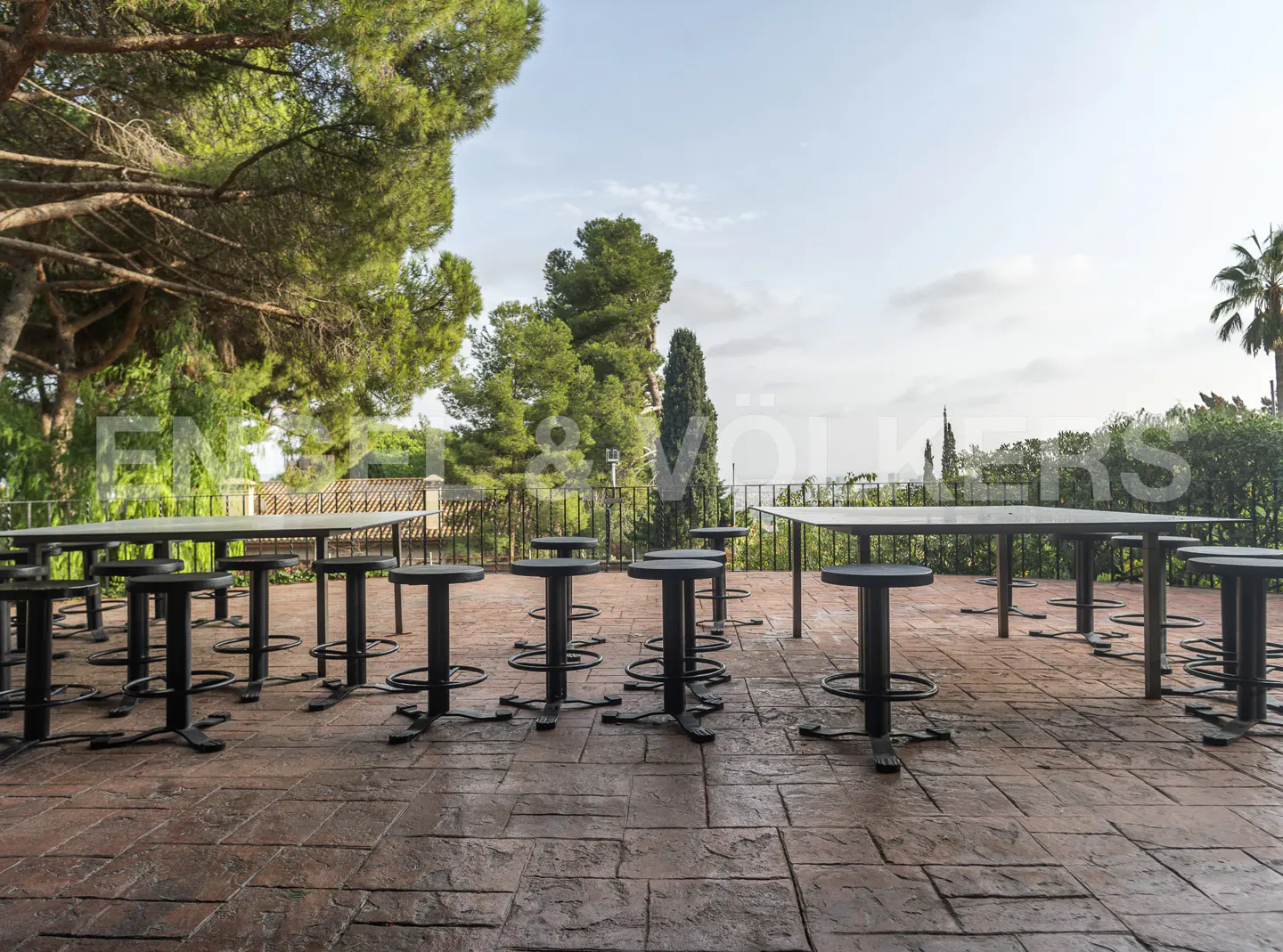 Outdoor patio with black stools and tables on a brick floor, with trees and sky in the background.