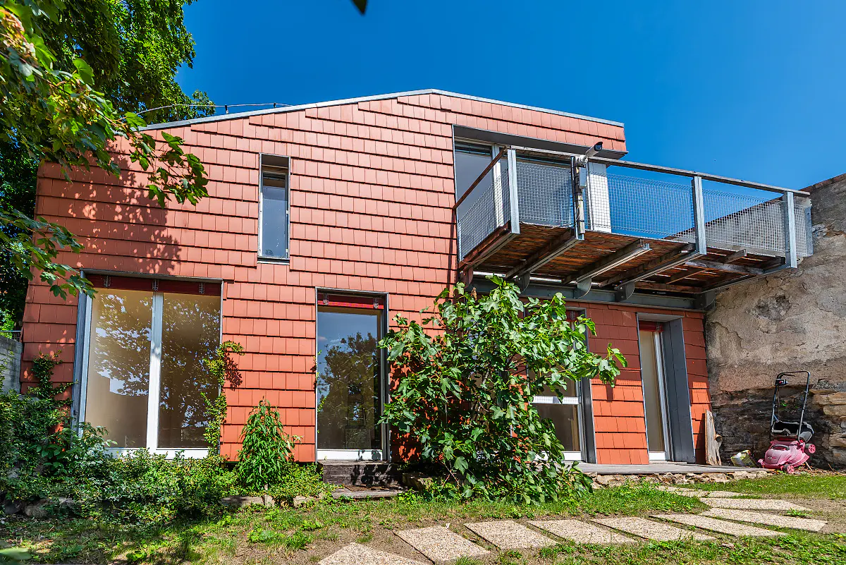 Modern two-story house with red tiled facade, a metal balcony, large glass doors, and a stone wall under a clear blue sky.
