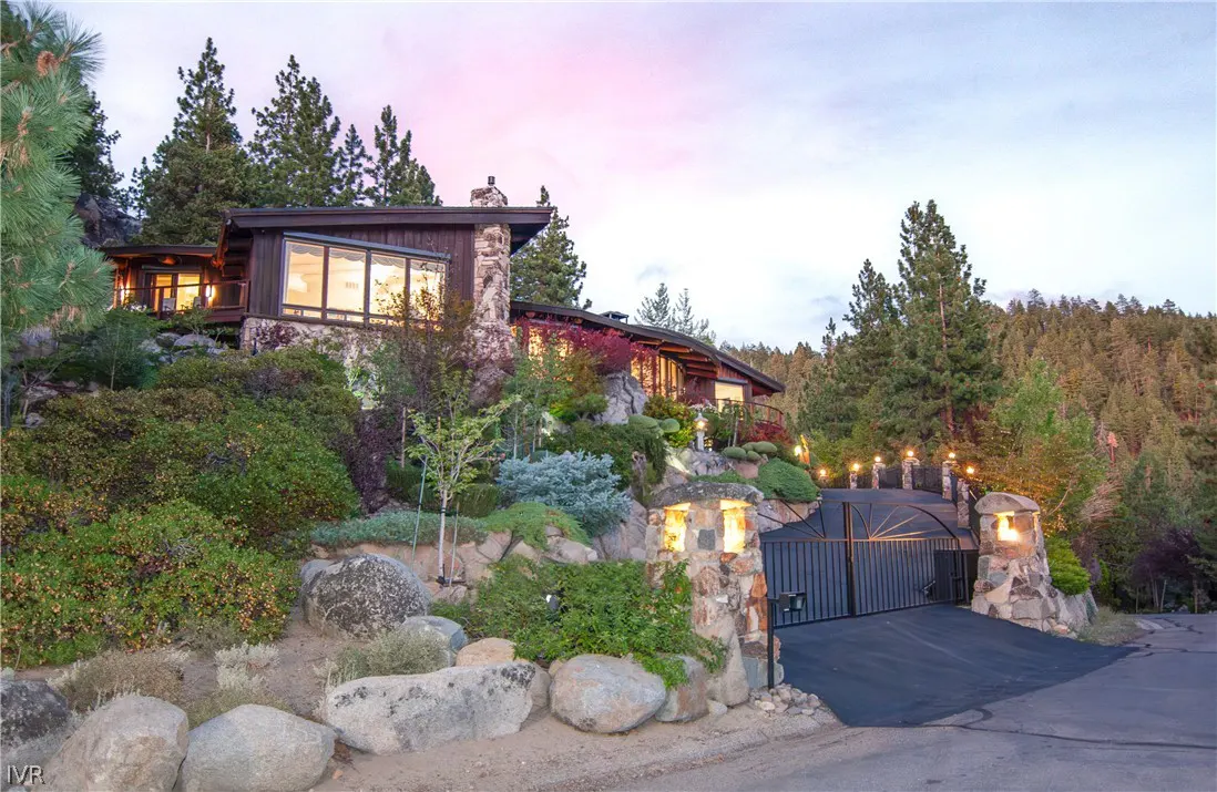 Modern mountain home with stone accents, large windows, and a gated driveway surrounded by lush landscaping and pine trees.