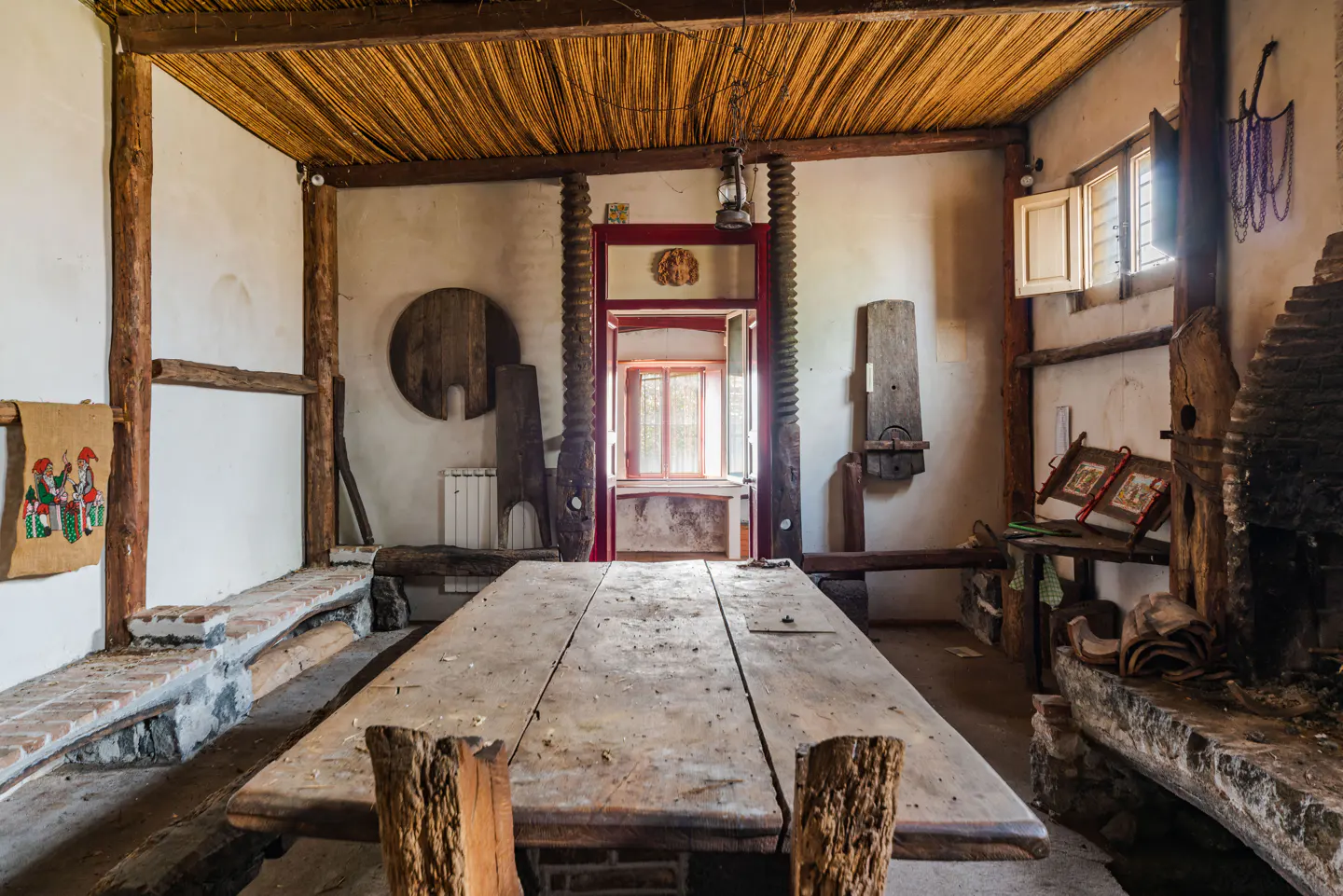 Rustic room with a long wooden table, straw ceiling, and stone benches. A red-framed doorway leads to another room.