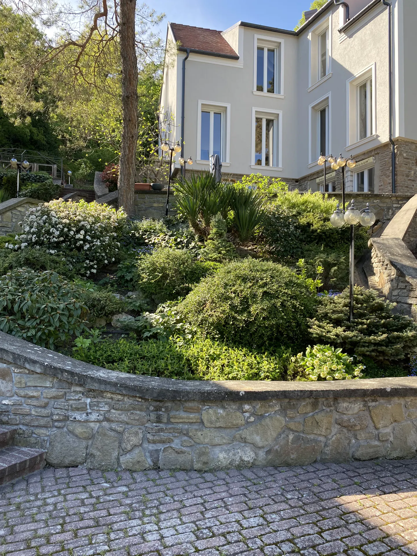 A gray two-story house with white trim is surrounded by lush green landscaping and stone walls.