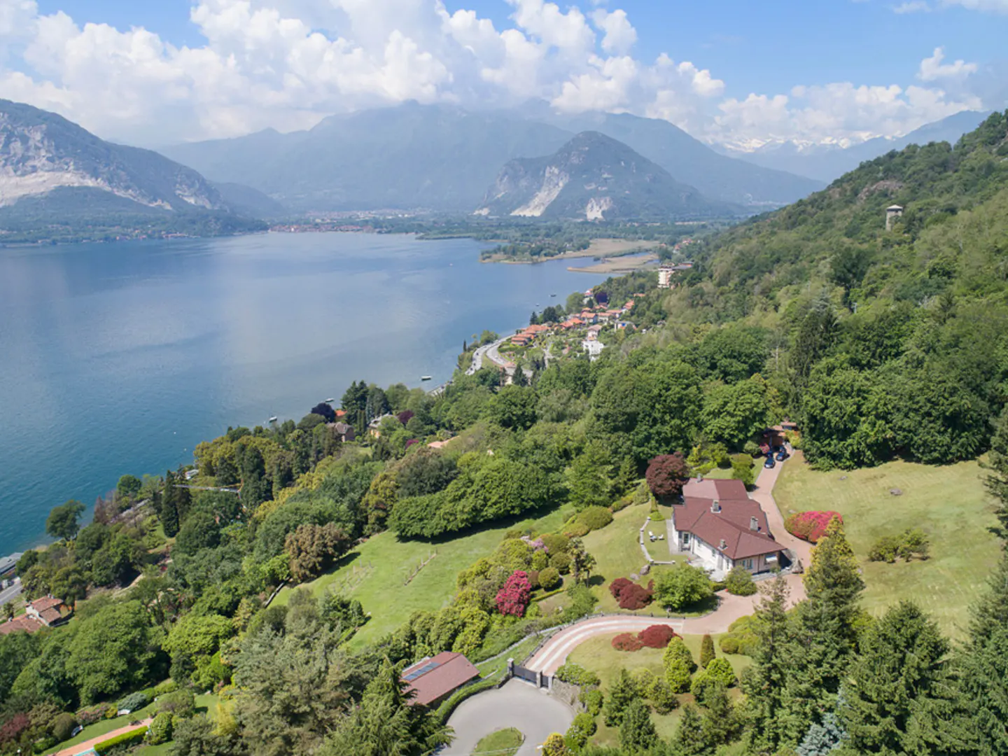 Aerial view of a large house with a red roof, surrounded by green trees, overlooking a blue lake and mountains.